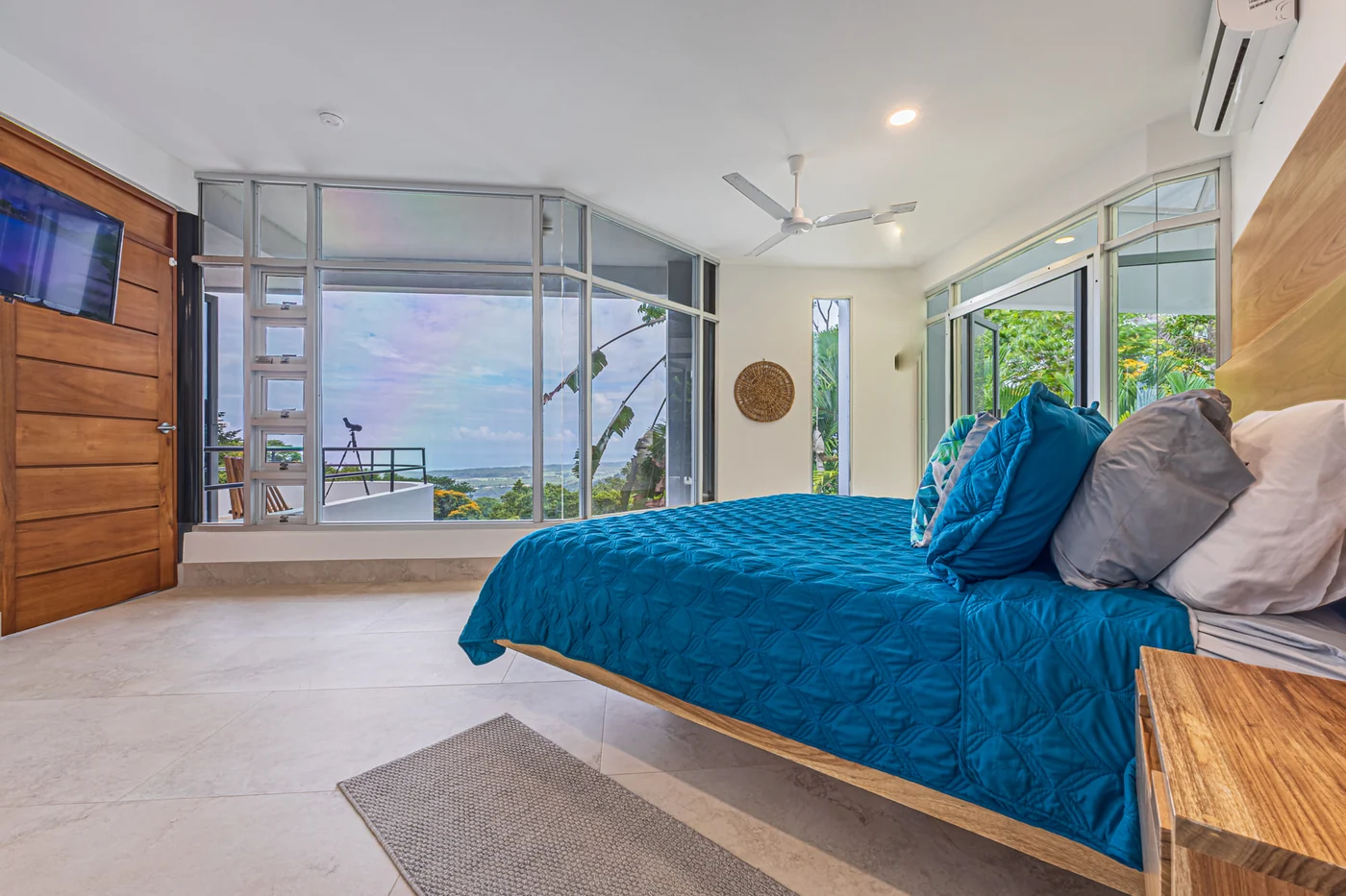 Habitación con cama king en ropa de cama turquesa y paredes de vidrio hacia la selva y el mar