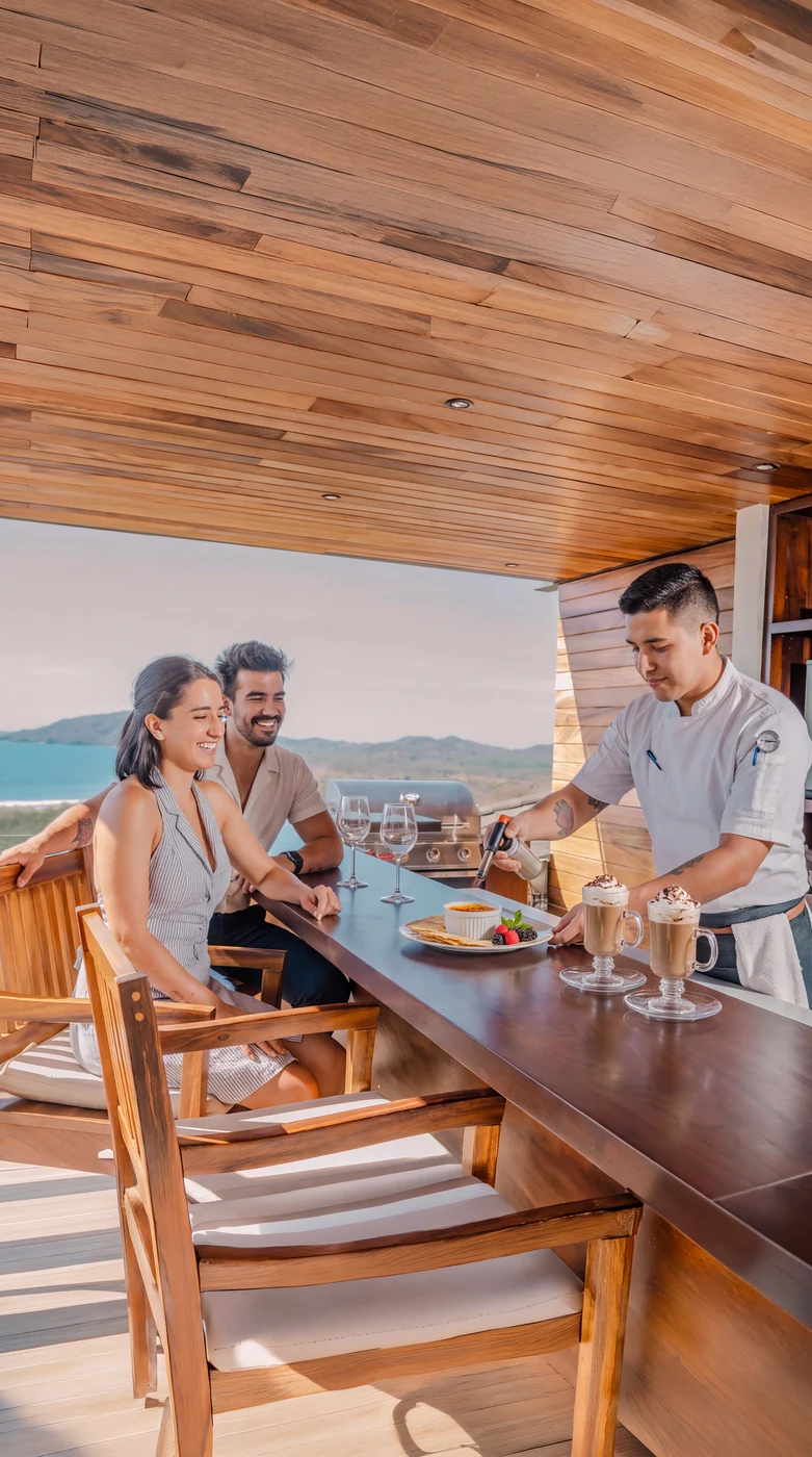 A private chef serving a couple at the dining table, the coastline through the windows behind